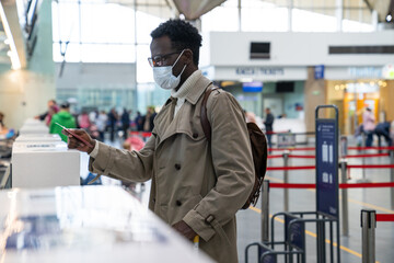 African American man stands at check-in counters at the airport terminal, giving passport to an officer. Flight rules during a Covid-19 pandemic only in a protective face mask. New normal concept.