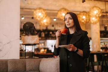 Beautiful young woman holding cup of tasty hot coffee in hand while standing in restaurant. Caucasian lady posing with glass of beverage in cafe with modern dark interior, looking away.