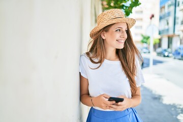 Young blonde woman on vacation smiling happy using smartphone leaning on the wall at street of city