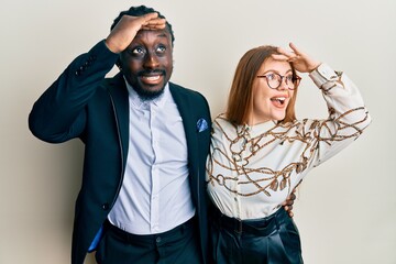 Young interracial couple wearing business and elegant clothes very happy and smiling looking far away with hand over head. searching concept.