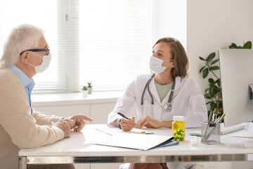 Young doctor in whitecoat and protective mask listening to senior patient