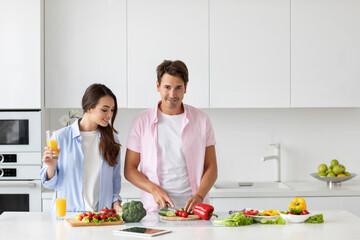 Handsome man cooking fresh vegetable salad, girl standing with a glass of orange juice in the kitchen.