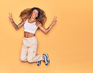 Young beautiful woman with tattoo wearing casual clothes smiling happy. Jumping with smile on face doing victory sign over isolated yellow background