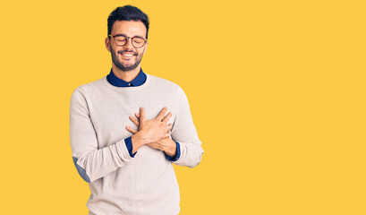 Young handsome hispanic man wearing elegant clothes and glasses smiling with hands on chest with closed eyes and grateful gesture on face. health concept.