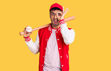 Young hispanic man playing baseball holding bat and ball smiling happy doing ok sign with hand on eye looking through fingers