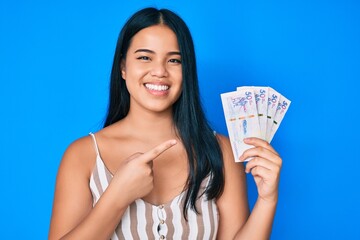 Young beautiful asian girl holding colombian pesos smiling happy pointing with hand and finger