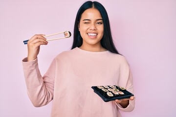 Young beautiful asian girl eating sushi using chopsticks winking looking at the camera with sexy...