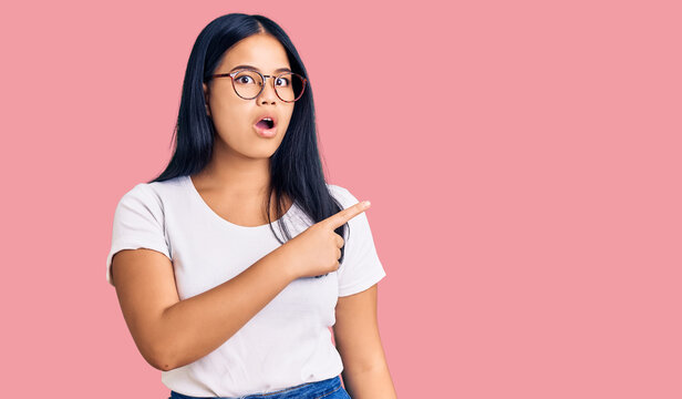 Young beautiful asian girl wearing casual clothes and glasses surprised pointing with finger to the side, open mouth amazed expression.