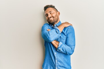 Handsome man with beard wearing casual denim jacket hugging oneself happy and positive, smiling...