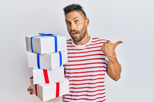 Handsome man with beard hodling with presents pointing thumb up to the side smiling happy with open mouth