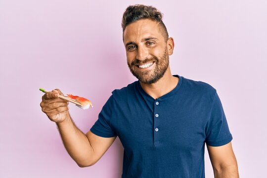 Handsome man with beard eating sushi using chopsticks looking positive and happy standing and smiling with a confident smile showing teeth