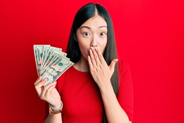 Young chinese woman holding 20 dollars banknote covering mouth with hand, shocked and afraid for mistake. surprised expression