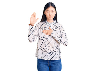 Young beautiful chinese woman wearing casual shirt swearing with hand on chest and open palm,...