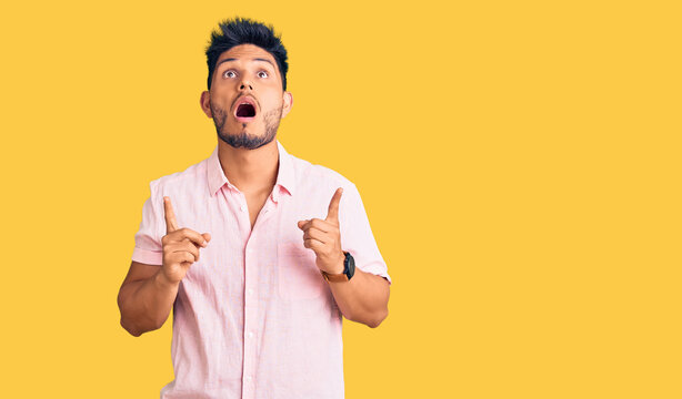 Handsome latin american young man wearing casual summer shirt amazed and surprised looking up and pointing with fingers and raised arms.