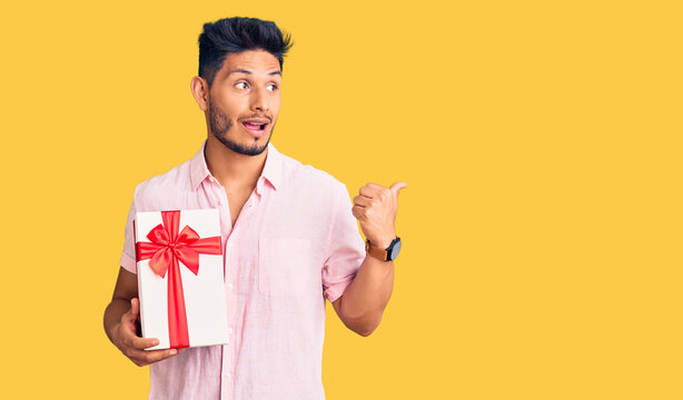 Handsome latin american young man holding gift pointing thumb up to the side smiling happy with open mouth