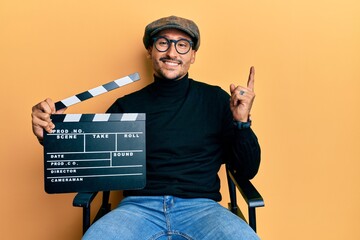 Handsome man with tattoos holding video film clapboard sitting on director chair smiling happy...