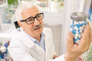 Obraz premium Aged drugstore worker in whitecoat and eyeglasses looking at big bottle of pills
