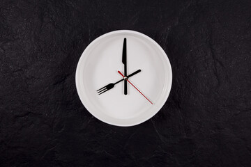 White plate clock on a dark textured stone background with copy space. The hands point to 8 o'clock. Concept - interval fasting or autophagy