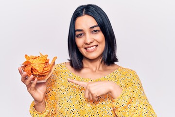 Young beautiful latin woman holding nachos potato chips smiling happy pointing with hand and finger