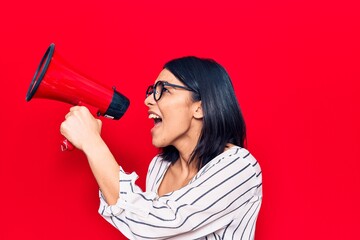 Young beautiful latin woman with angry expression. Screaming loud using megaphone standing over isolated red background