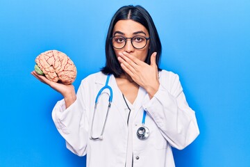 Young beautiful latin woman wearing doctor stethoscope holding brain covering mouth with hand,...
