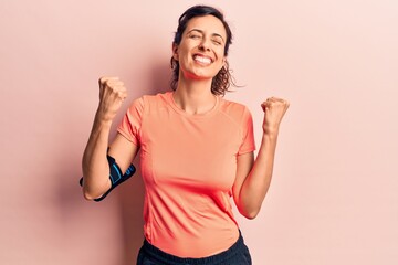 Young beautiful hispanic woman wearing sportswear using smartphone very happy and excited doing winner gesture with arms raised, smiling and screaming for success. celebration concept.