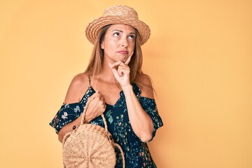Middle age hispanic woman wearing summer hat holding wicker bag serious face thinking about...