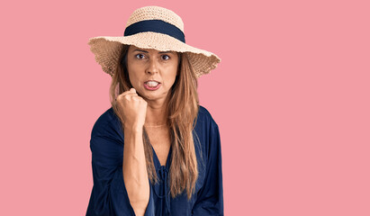 Middle age hispanic woman wearing summer hat angry and mad raising fist frustrated and furious while shouting with anger. rage and aggressive concept.