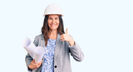 Young beautiful brunette woman wearing architect hardhat holding blueprint smiling happy and positive, thumb up doing excellent and approval sign