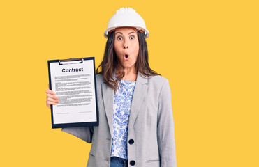 Young beautiful brunette woman wearing architect hardhat holding contract scared and amazed with open mouth for surprise, disbelief face