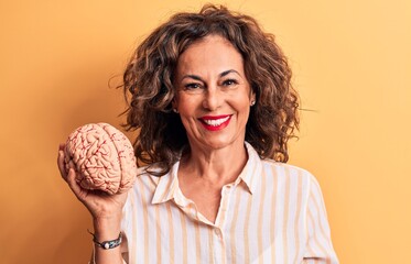 Middle age brunette woman asking for care memory holding brain over yellow background looking...
