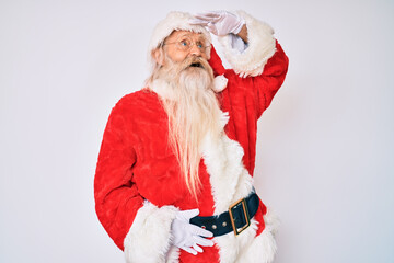 Old senior man with grey hair and long beard wearing santa claus costume with suspenders very happy and smiling looking far away with hand over head. searching concept.