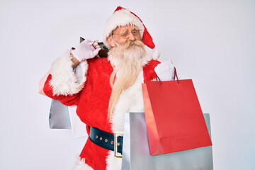 Old senior man with grey hair and long beard wearing santa claus costume holding shopping bags winking looking at the camera with sexy expression, cheerful and happy face.