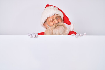 Old senior man with grey hair and long beard wearing santa claus costume holding banner winking...