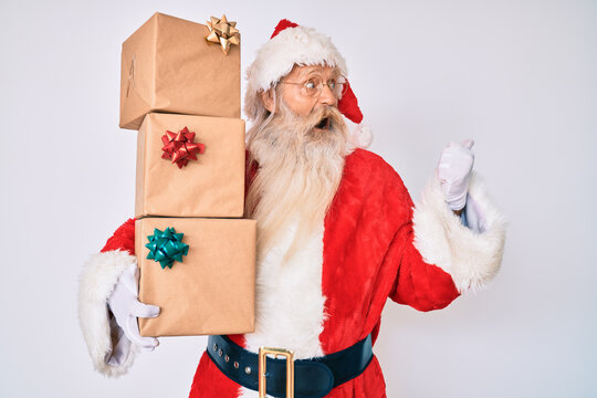 Old senior man with grey hair and long beard wearing santa claus costume holding presents pointing thumb up to the side smiling happy with open mouth