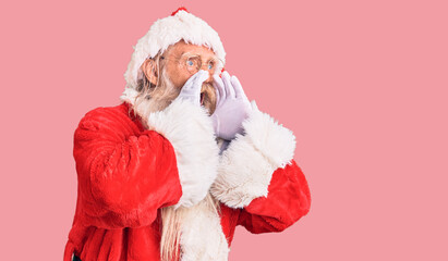 Old senior man with grey hair and long beard wearing traditional santa claus costume shouting angry out loud with hands over mouth
