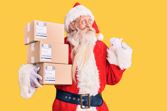 Old senior man with grey hair and long beard wearing santa claus costume holding boxes pointing thumb up to the side smiling happy with open mouth