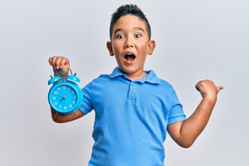 Little boy hispanic kid holding alarm clock pointing thumb up to the side smiling happy with open...