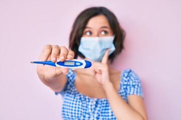Young beautiful woman wearing medical mask holding holding thermometer serious face thinking about question with hand on chin, thoughtful about confusing idea