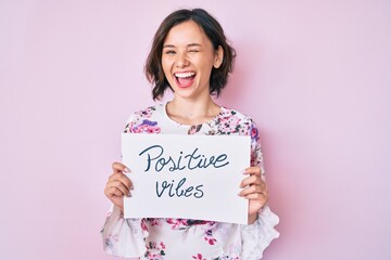 Young beautiful girl holding positive vibes banner winking looking at the camera with sexy...