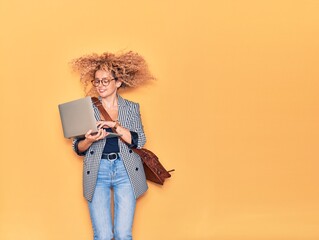 Young beautiful curly businesswoman wearing leather bag smiling happy. Jumping with smile on face working using laptop over isolated yellow background.