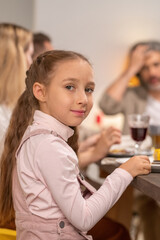 Cute girl with long brown hair looking at you while sitting by festive table