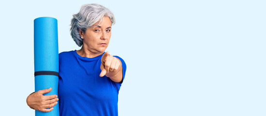 Senior woman with gray hair holding yoga mat pointing with finger to the camera and to you, confident gesture looking serious - obrazy, fototapety, plakaty