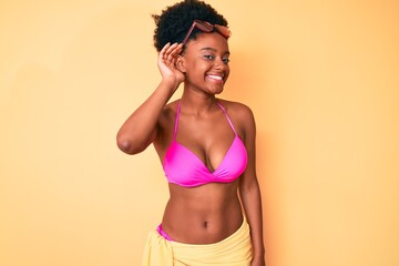 Naklejka premium Young african american woman wearing bikini smiling with hand over ear listening an hearing to rumor or gossip. deafness concept.