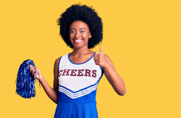 Young african american woman wearing cheerleader uniform holding pompom doing happy thumbs up gesture with hand. approving expression looking at the camera showing success.