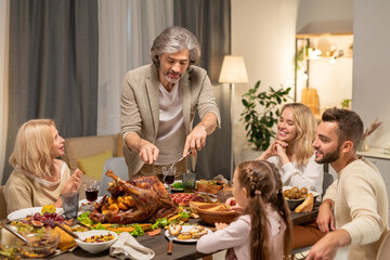 Aged man with knife and fork cutting homemade roasted turkey by family dinner
