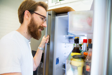 Naklejka na ściany i meble Young man looking into an open home fridge, a refrigerator.