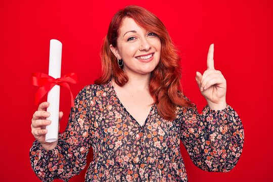 Young beautiful redhead woman holding graduated degree diploma over red background smiling happy pointing with hand and finger to the side