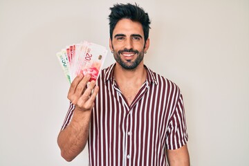 Handsome hispanic man with beard holding israel shekels looking positive and happy standing and...