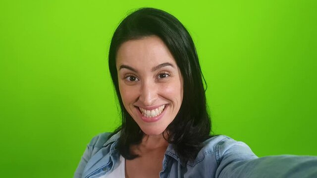 smiling woman posing on green screen background. Girl taking selfie self portrait photo on smartphone. Chroma key. Female model showing positive face emotions
